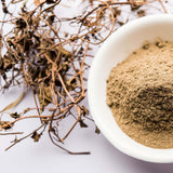 White bowl filled with Bhringraj powder next to dried herbs on a light background