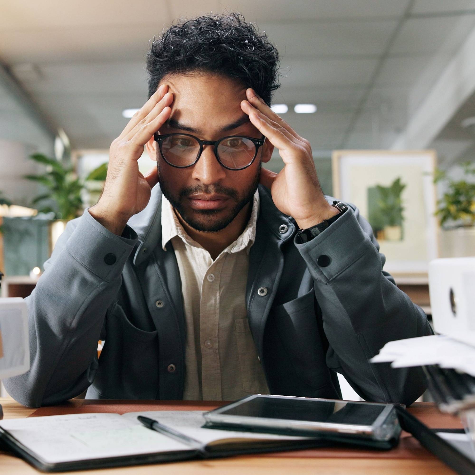 Man sitting at a desk with a tablet and papers, appearing stressed.