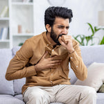 Man sitting on a couch with a concerned expression, holding his chest and coughing