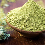 Ayumi Henna powder in a wooden bowl on a wooden surface with green leaves.