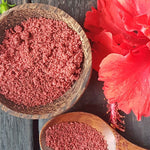 Wooden bowl and spoon filled with Hibiscus powder next to a red flower on a dark surface