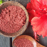 Wooden bowl and spoon filled with Hibiscus powder next to a red flower on a dark surface