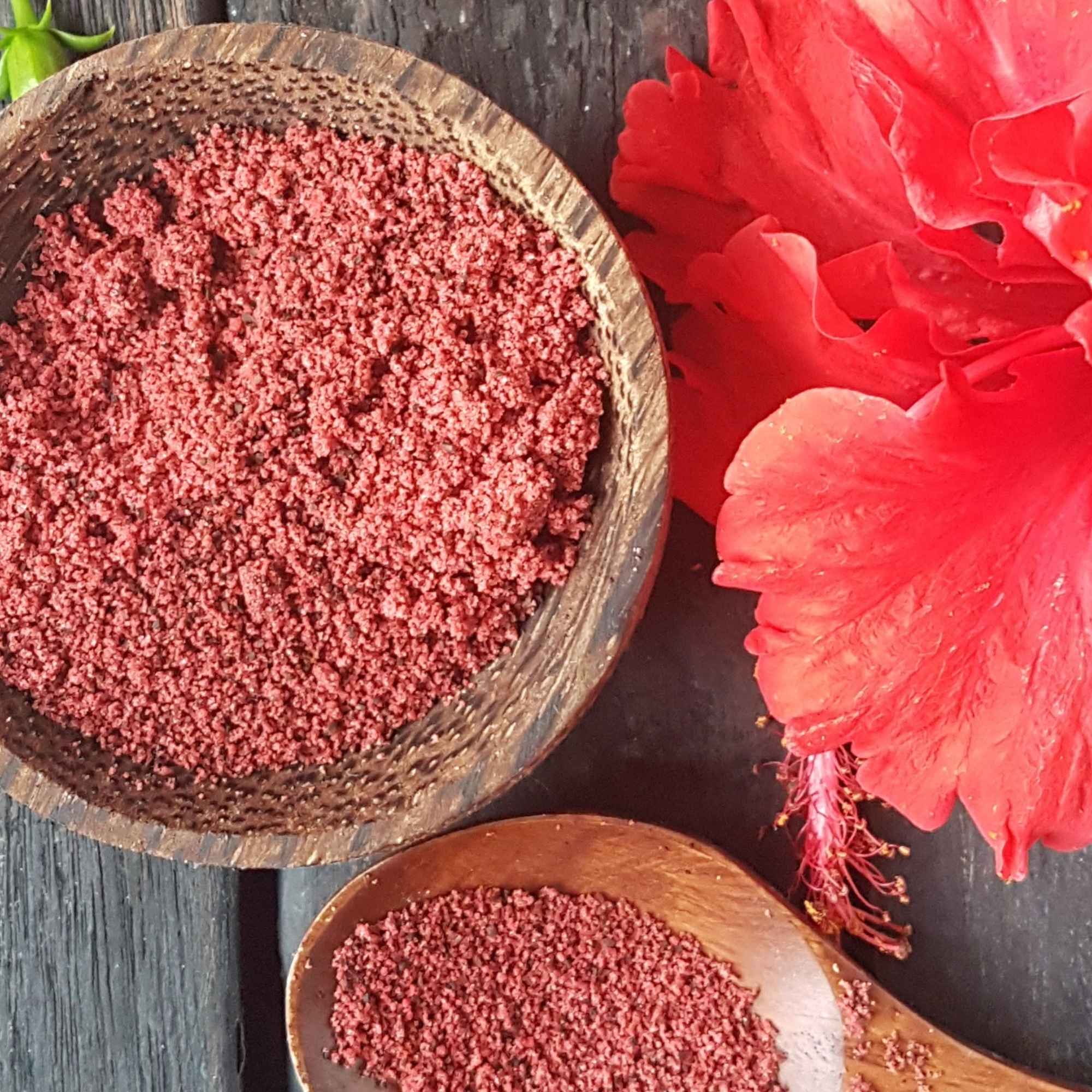 Wooden bowl and spoon filled with Hibiscus powder next to a red flower on a dark surface