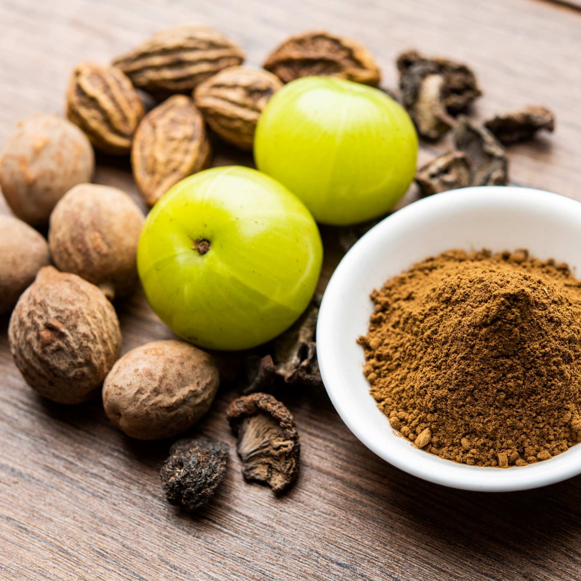 Raw Ingredients of Triphala, Amla, Beda and Harde, and a bowl of ground Tripohala on a wooden surface.