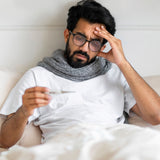 Man with a beard wearing glasses and a scarf, holding a thermometer, in a white room.