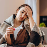 Woman wrapped in a blanket, holding a thermometer, looking unwell.