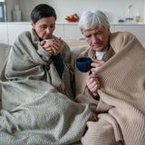 Two people sitting on a couch, wrapped in blankets and holding mugs, in a home setting.