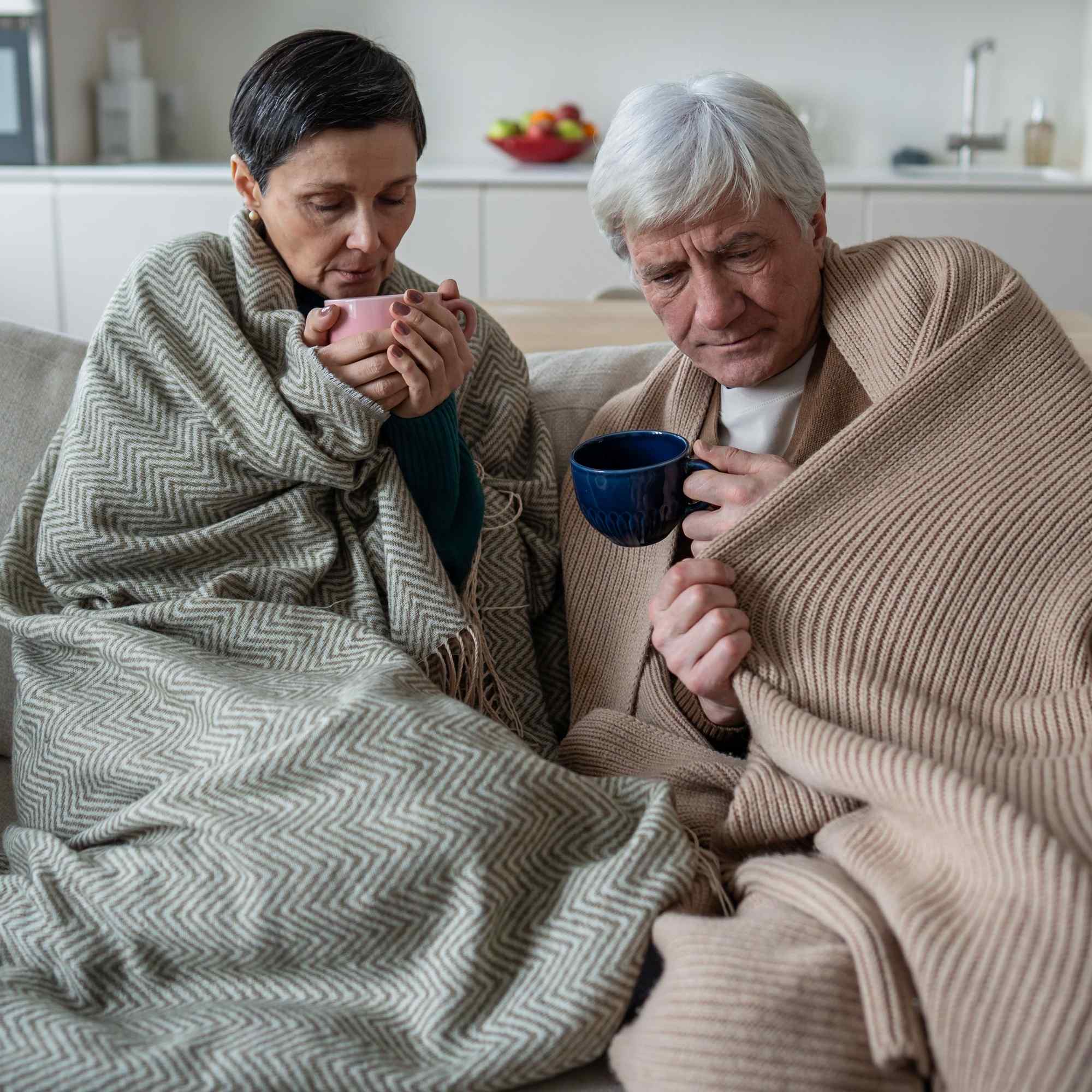 Two people sitting on a couch, wrapped in blankets and holding mugs, in a home setting.