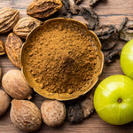 Amla powder in a gold bowl with raw ingredients, Amla, Beda & Harde surrounding 