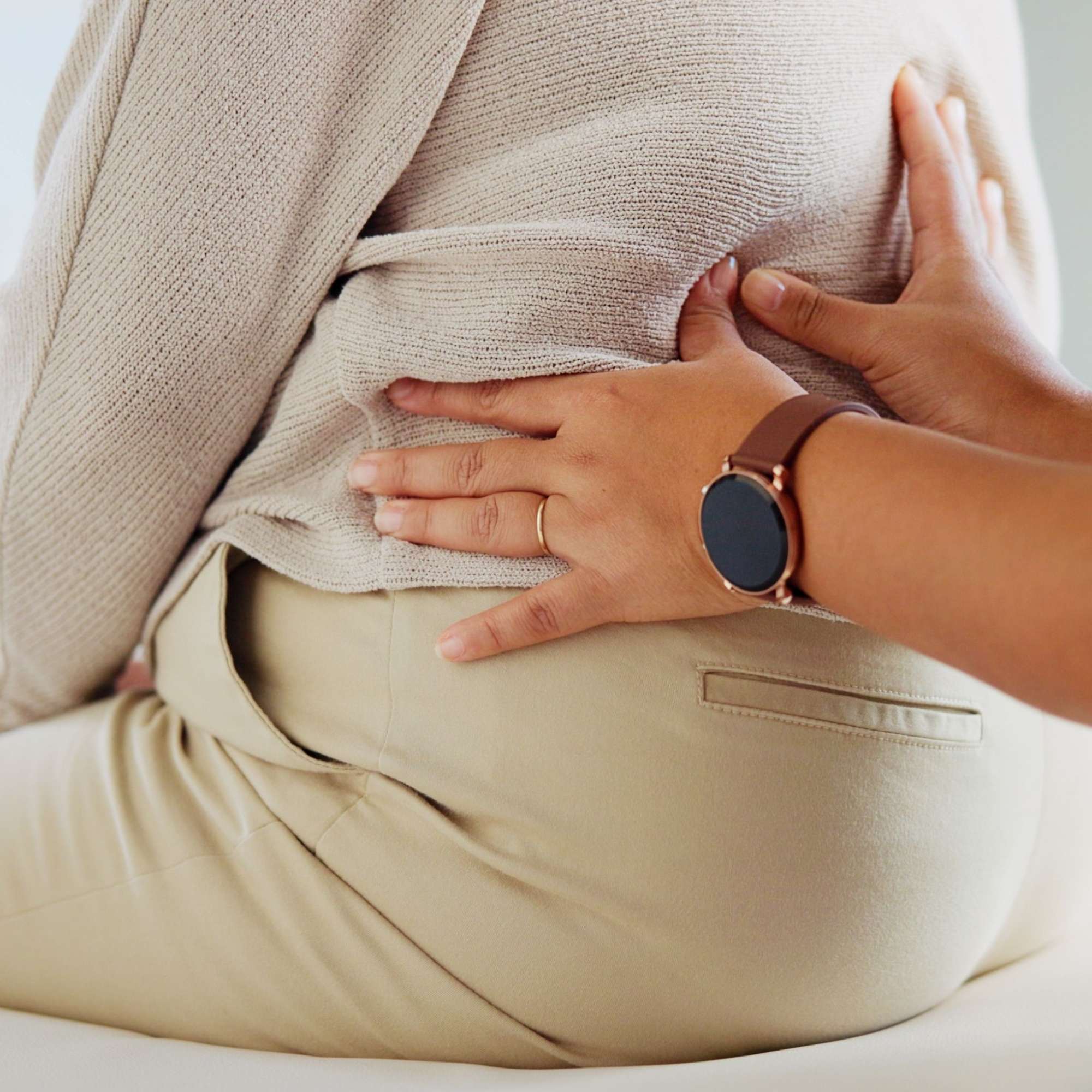 Person wearing a beige sweater and pants with hands on their stomach, wearing a watch.